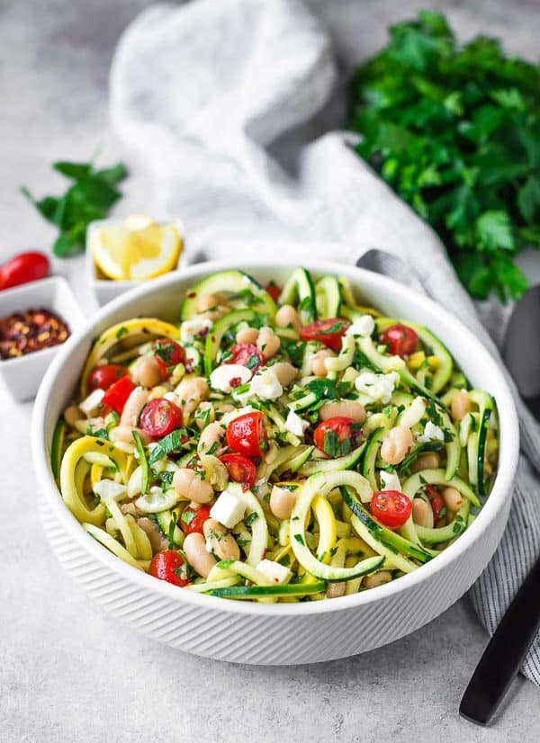 A white bowl full of zucchini and summer squash noodles, feta, great northern beans, tomatoes, and fresh herbs.