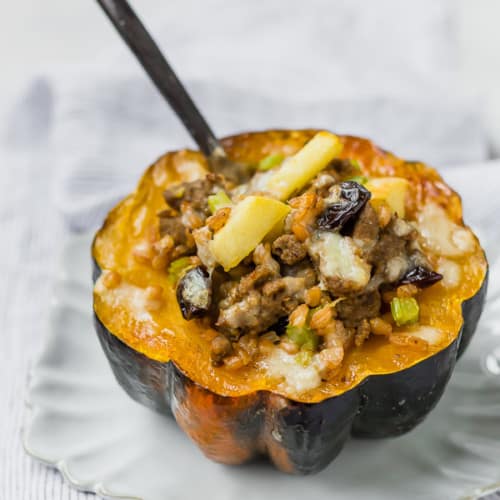 Acorn squash half on a white plate. Squash is filled with farro, sausage, apples, cranberries, and cheese.