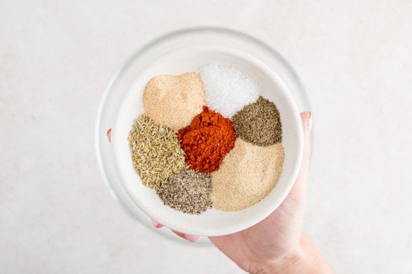 A hand holding a bowl filled with various spices and herbs for stew seasoning.