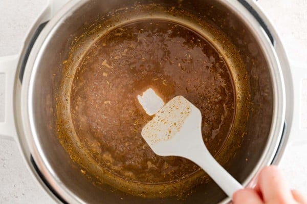 A plastic spatula is used to scrape the stuck-on bits from the bottom of the Instant Pot after deglazing with broth.