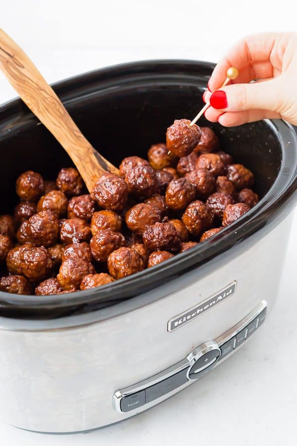 Meatballs in slow cooker, with hand skewering a meatball on a toothpick.