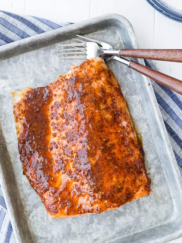 Overhead view of baked maple salmon on a sheet pan, next to a pair of forks with a blue striped towel in background.