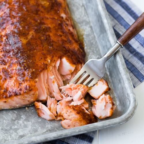 Close up view of baked maple salmon on a sheet pan, flaked with a fork with a blue striped towel in background.