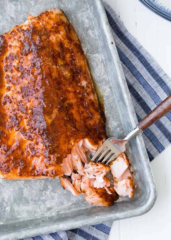 Overhead view of baked maple salmon on a sheet pan, flaked with a fork with a blue striped towel in background.