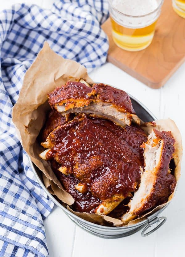 Overhead of bbq ribs in metal basket with brown paper, blue plaid cloth nearby along with glass of beer.