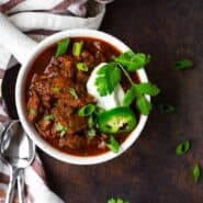 image of texas style chili in a white bowl with a handle, garnished with sour cream, cilantro, and jalapeno