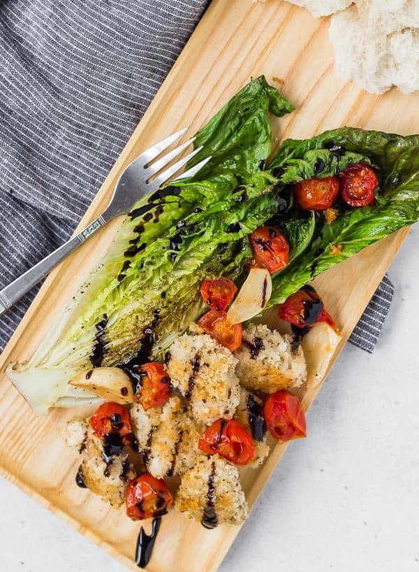 Image of Roasted Romaine salad with roasted tomatoes, roasted garlic, crispy chicken and balsamic glaze. Served on a wooden tray with a fork. 