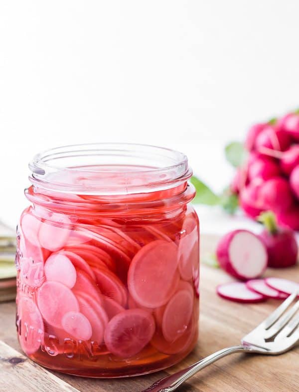 Pickled sliced radishes in open jar with fork. A bunch of fresh radishes in background.