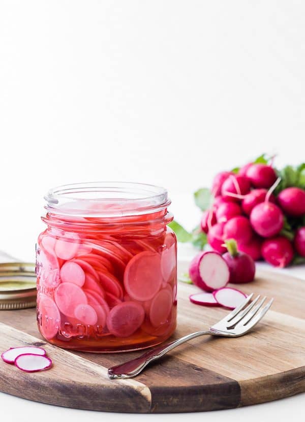 Jar of pickled radishes, on wooden cutting board along with fresh radishes.