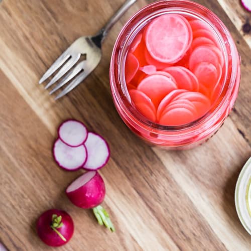 Overhead of open jar of pickled radishes on wooden cutting board, with fork and fresh radishes.