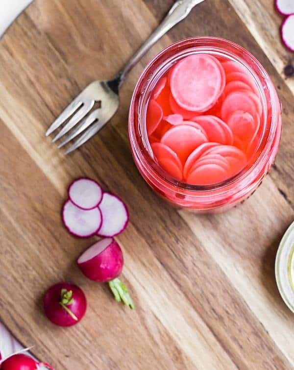 Overhead of open jar of pickled radishes with fork, and sliced radishes along side.