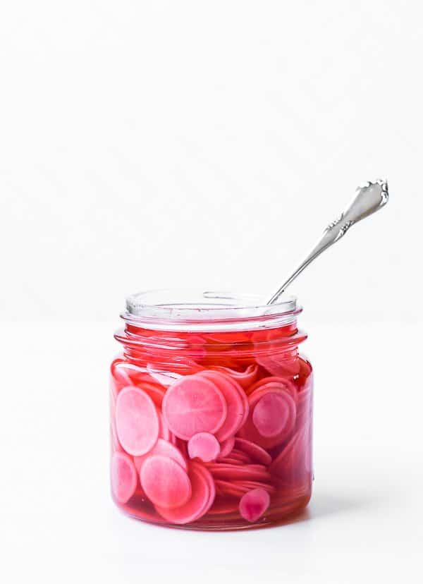 Jar of pickled radishes in open jar with fork inserted, on totally white background.