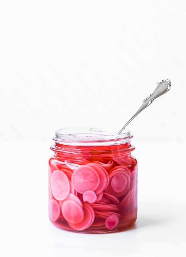 Jar of pickled radishes in open jar with fork inserted, on totally white background.