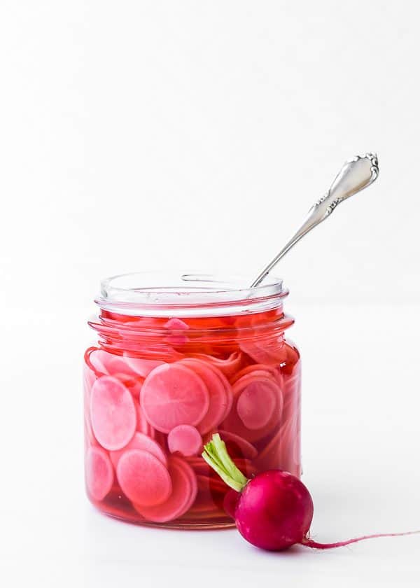 image of jar of pickled radishes - quick pickled radish recipe. One whole radish sits in front. Bright white background.