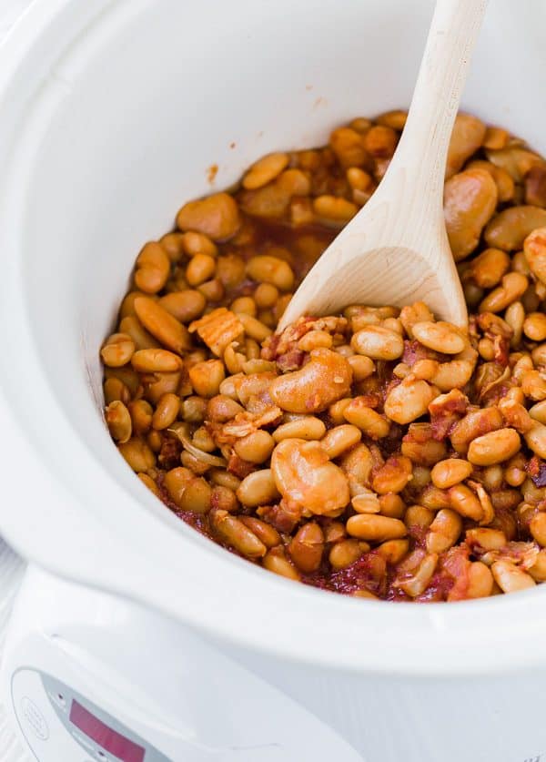 image of slow cooker baked beans in a white slow cooker with a wooden spoon