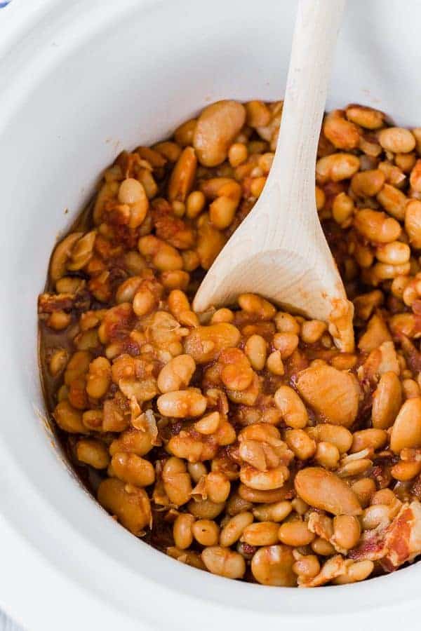 Image of crock pot baked beans in a white slow cooker with a wooden spoon stuck in the middle.