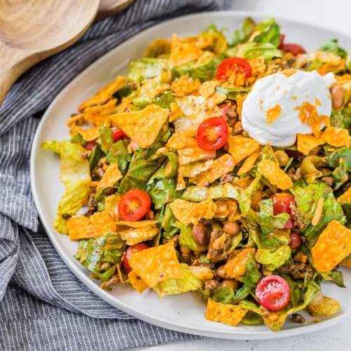 image of a classic taco salad recipe on a plate with a black and white linen and wooden serving utensils in the background of the photo.