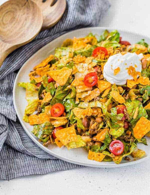 image of a classic taco salad recipe on a plate with a black and white linen and wooden serving utensils in the background of the photo.