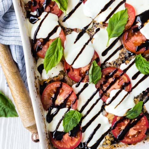 overhead image of caprese quinoa bake, sprinkled with fresh basil and drizzled with balsamic reduction. Wooden spoon and linen next to white baking dish.