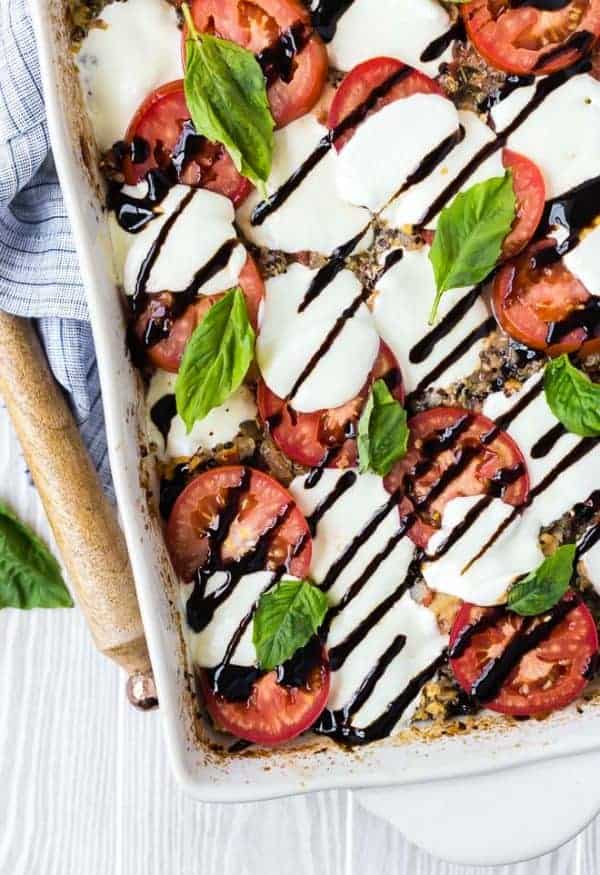 overhead image of caprese quinoa bake, sprinkled with fresh basil and drizzled with balsamic reduction. Wooden spoon and linen next to white baking dish.