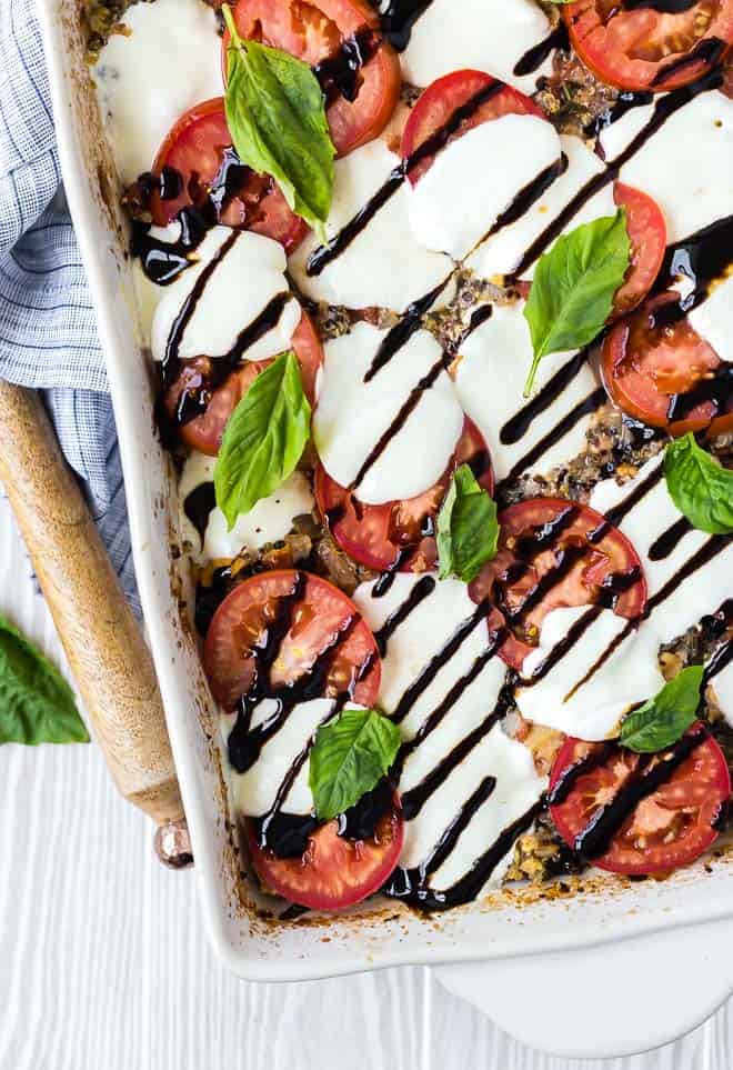 Made in one pan and largely hands-off, this chicken caprese quinoa bake is a vibrant taste of summer with very little work! overhead image of caprese quinoa bake, sprinkled with fresh basil and drizzled with balsamic reduction. Wooden spoon and linen next to white baking dish.