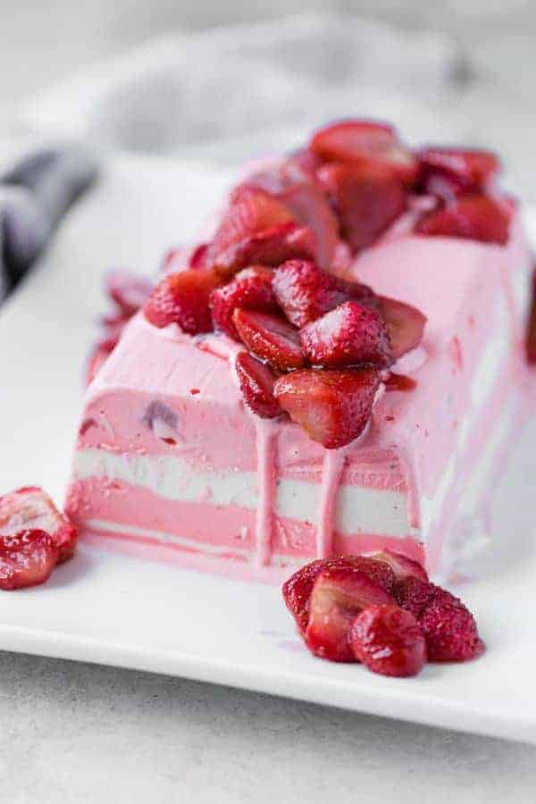 Image of layered strawberry and vanilla ice cream terrine topped with roasted strawberries. Arranged on a white rectangular plate.