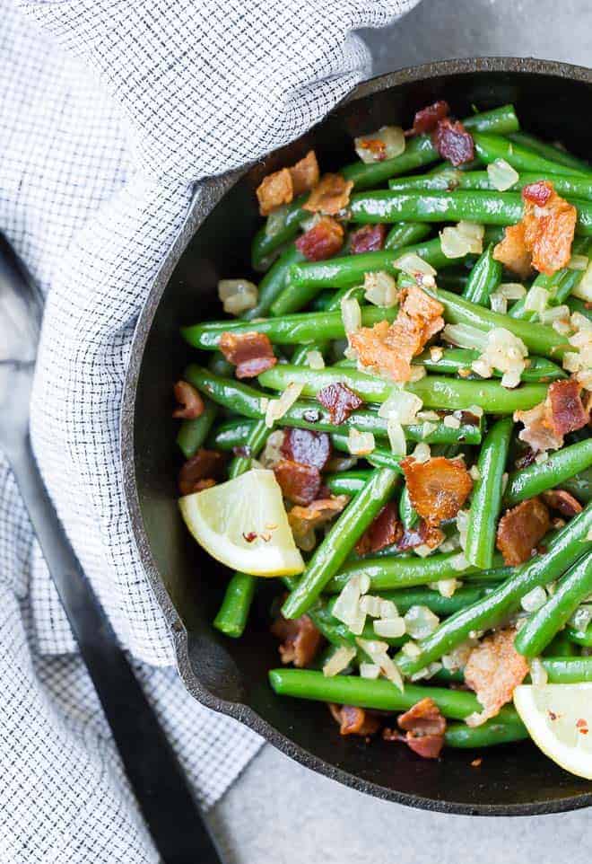 Image of vibrant green beans in a black cast iron skillet, sprinkled with garlic, onion, and bacon. Lemon wedges are used for garnish.