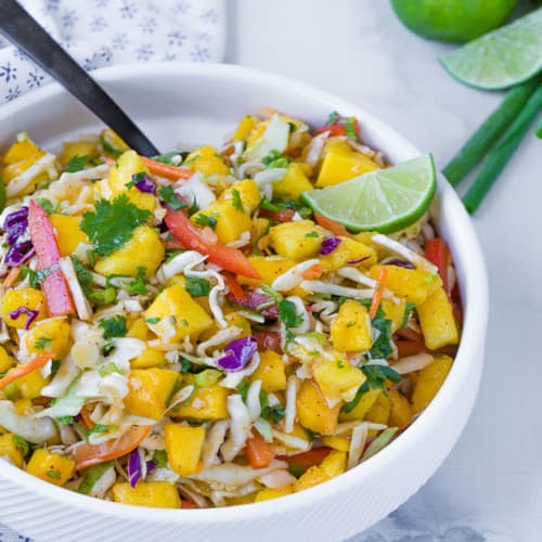 image of bright and colorful mango slaw, the perfect slaw for fish tacos, in a white bowl with a black spoon. Garnished with a lime.