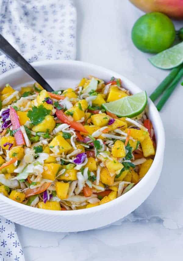 image of bright and colorful mango slaw, the perfect slaw for fish tacos, in a white bowl with a black spoon. Garnished with a lime. 