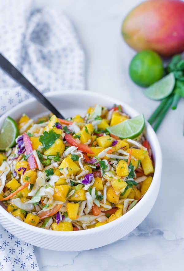 Image of mango slaw for tacos, in a white bowl with a mango and limes in the background