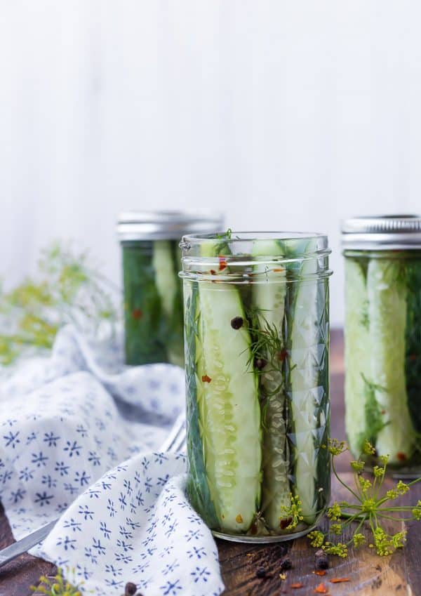 Image of quick dill pickles in a jar. Two pickle bars in the background are also pictured. 