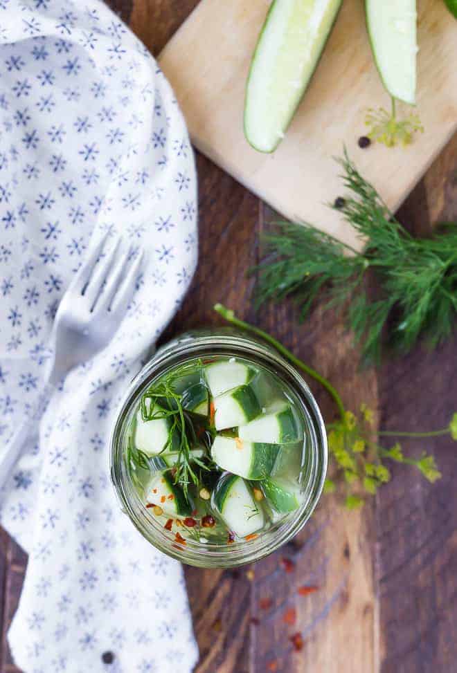 Overhead of pickles in jar, with fork, cloth, fresh dill and sliced cucumbers arranged around.