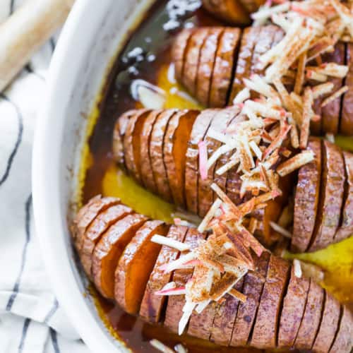 Four hasselback sweet potatoes in a white baking dish, topped with apples.