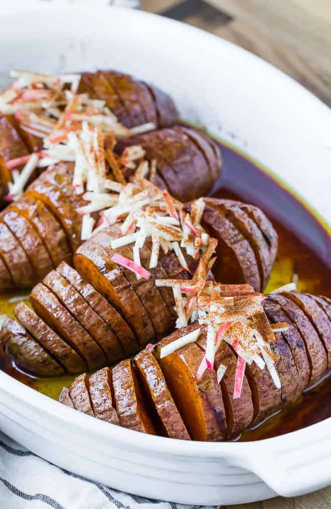 Four hasselback sweet potatoes in a white baking dish, topped with apples.