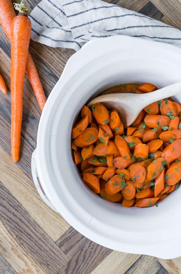 Crockpot glazed carrots in a white slow cooker, with a wooden spoon. Parsley is sprinkled on top.