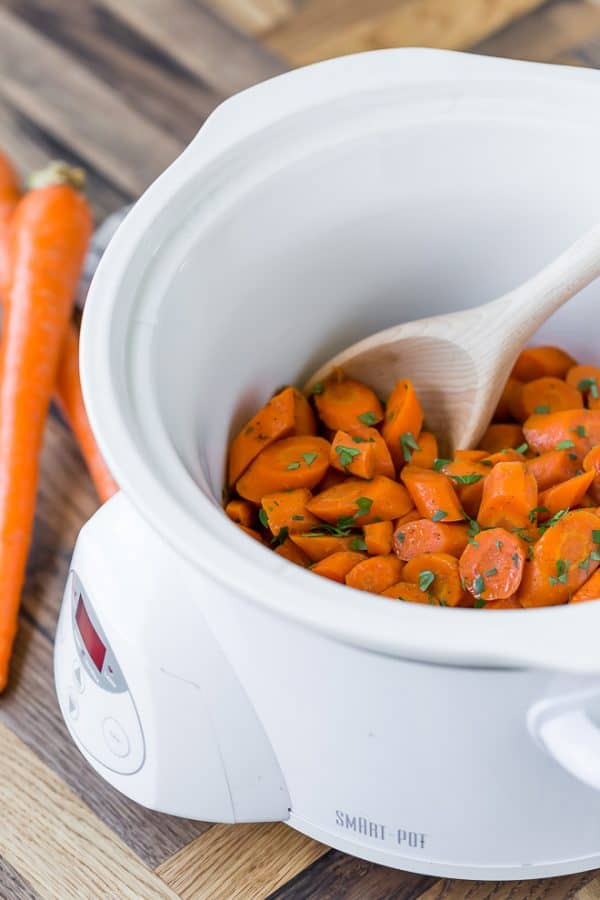 Slow cooker glazed carrots in a white slow cooker with a wooden spoon..