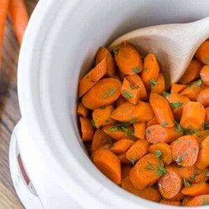 Crockpot glazed carrots in a white slow cooker, with a wooden spoon. Parsley is sprinkled on top.