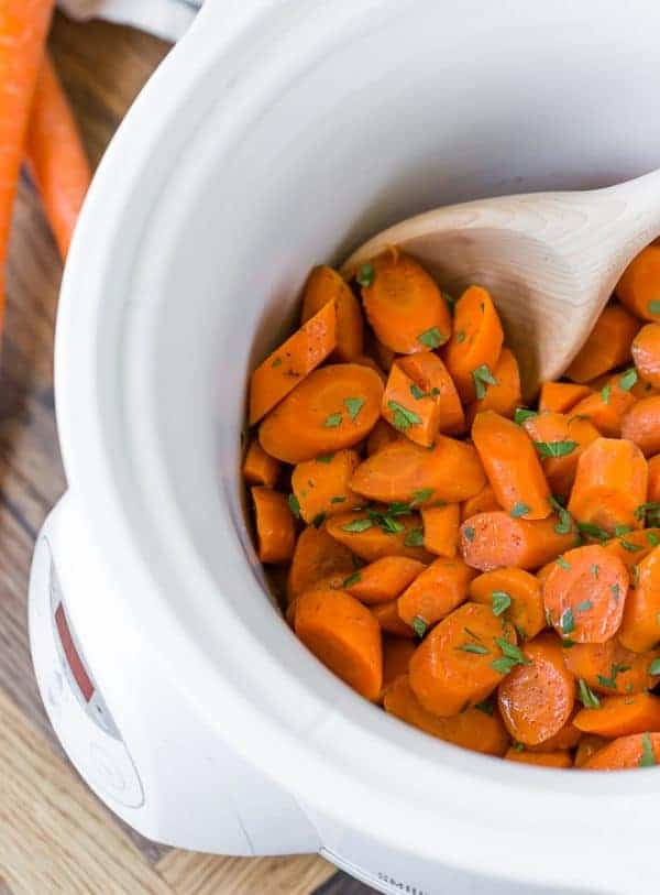 Crockpot glazed carrots in a white slow cooker, with a wooden spoon. Parsley is sprinkled on top.