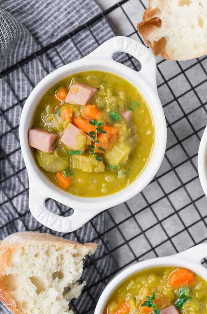 Image of split pea soup recipe with ham, carrots, and thyme. In a small white crock with bread next to it.