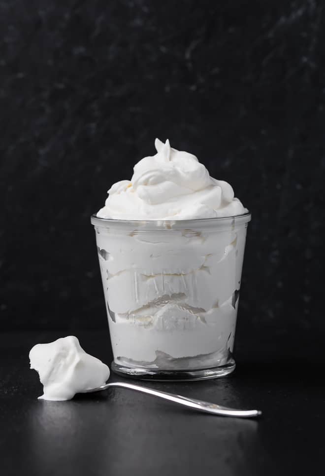 Image of freshly whipped whipped cream in a glass. A spoonful of whipped cream sits on the surface next to it. Photograph taken on a black background.
