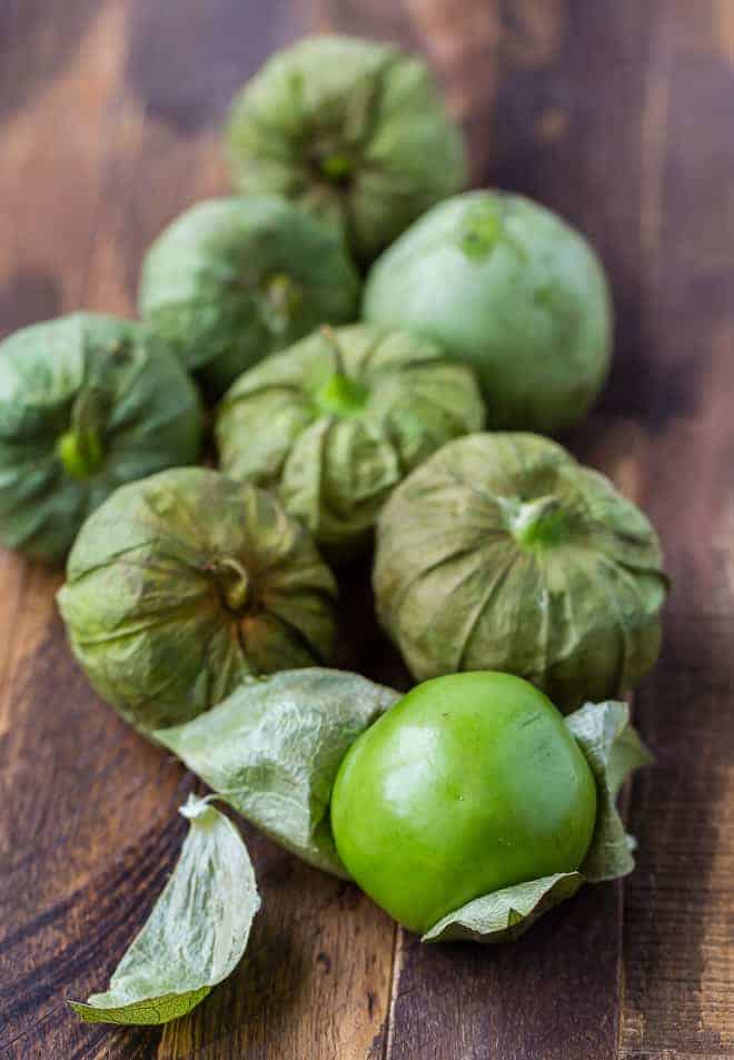 Image of tomatillos on a wooden surface. The tomatillo in the foreground has its husks peeled away.