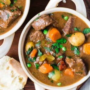Image of crockpot beef stew in a white bowl with two handles. The beef stew is garnished with parsley and served with bread.