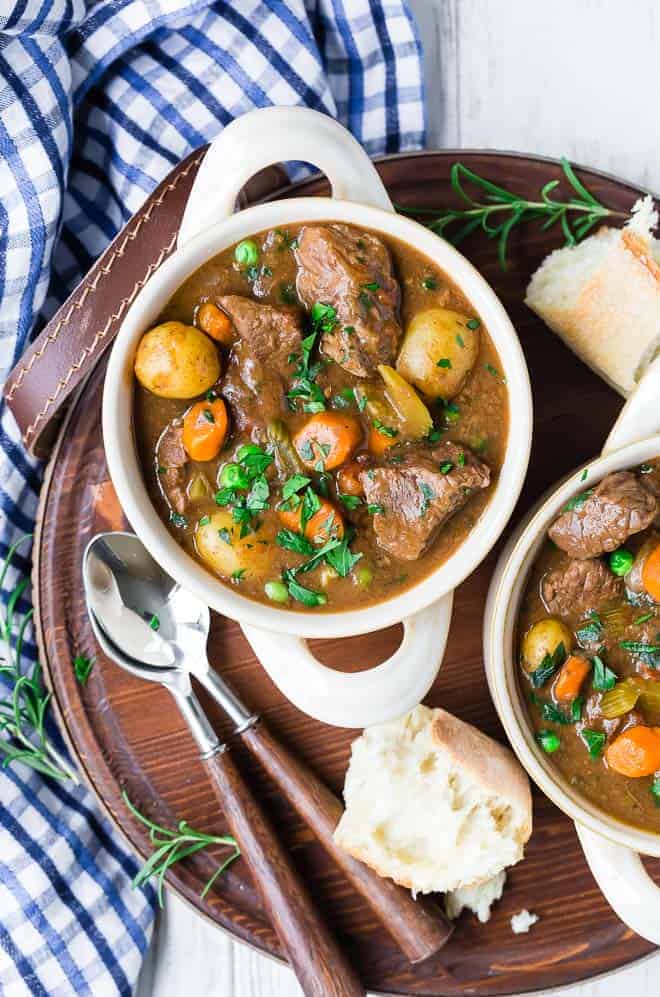 Photograph of a bowl of hearty beef stew that's been made in a crockpot. Two bowls are pictured, along with two spoons and a few pieces of bread.