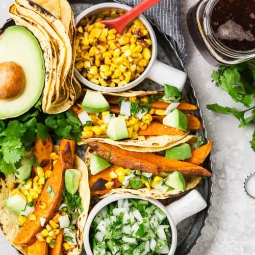 Image of vegan sweet potato tacos on a platter with charred corn, onions and cilantro, and avocado. A glass of beer sits next to the platter.