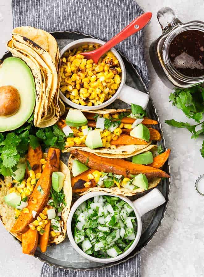 Image of vegan sweet potato tacos on a platter with charred corn, onions and cilantro, and avocado. A glass of beer sits next to the platter.