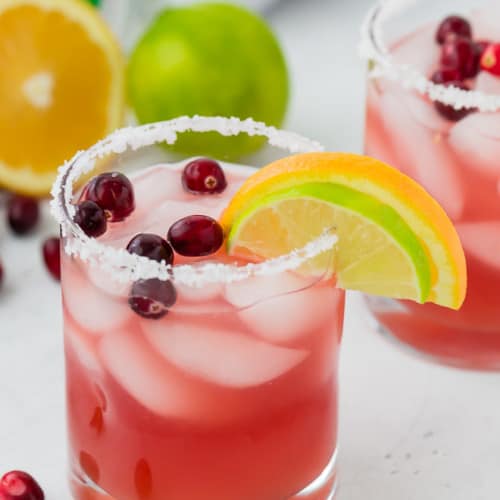 Image of a beautiful cranberry margarita in a salt rimmed glass, garnished with a lime wedge, an orange wedge, and fresh cranberries.