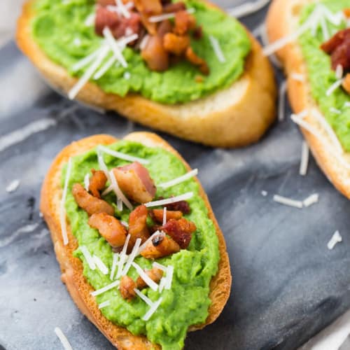 Image of pea crostini on a black marble serving platter, topped with pancetta and parmesan.