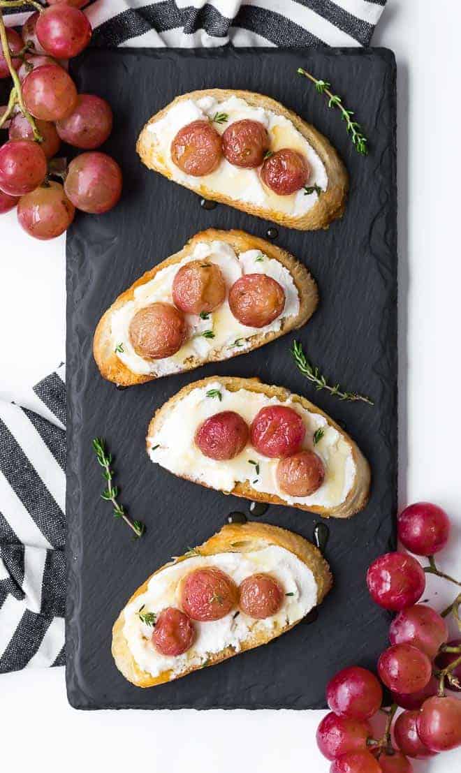 Image of four crostini on a small black slate board. Crostini is topped with roasted grapes and ricotta cheese.