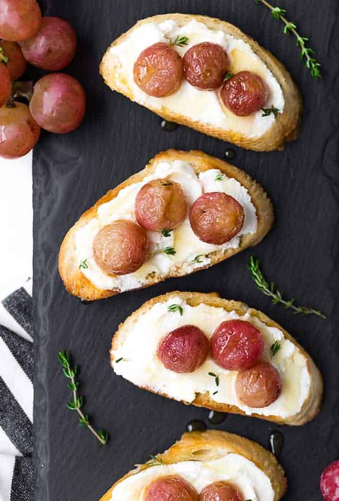 Image of roasted grape crostini on a black slate background. Fresh thyme is sprinkled around them.