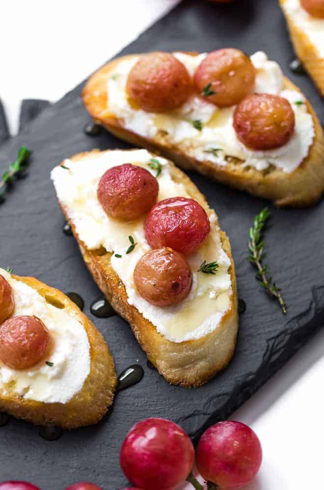 Image of crostini on a black slate board. It is stopped with ricotta cheese, roasted grapes, honey, and thyme.